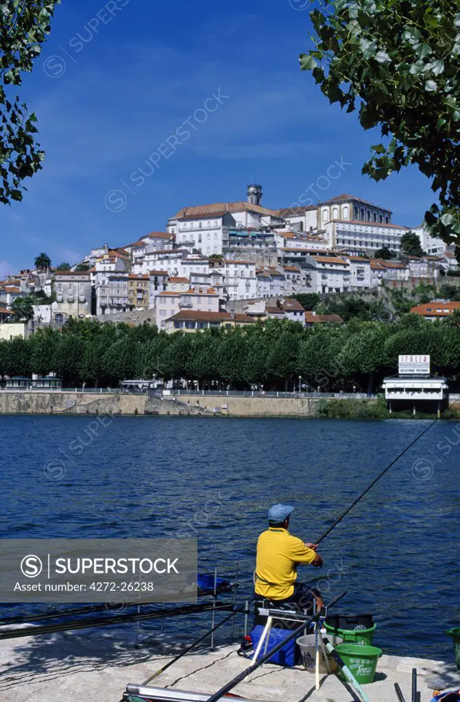 A fisherman sits on the bank of the Rio Mondego looking towards Coimbra with the university on the summit of the hill and the tightly packed houses of the old town below