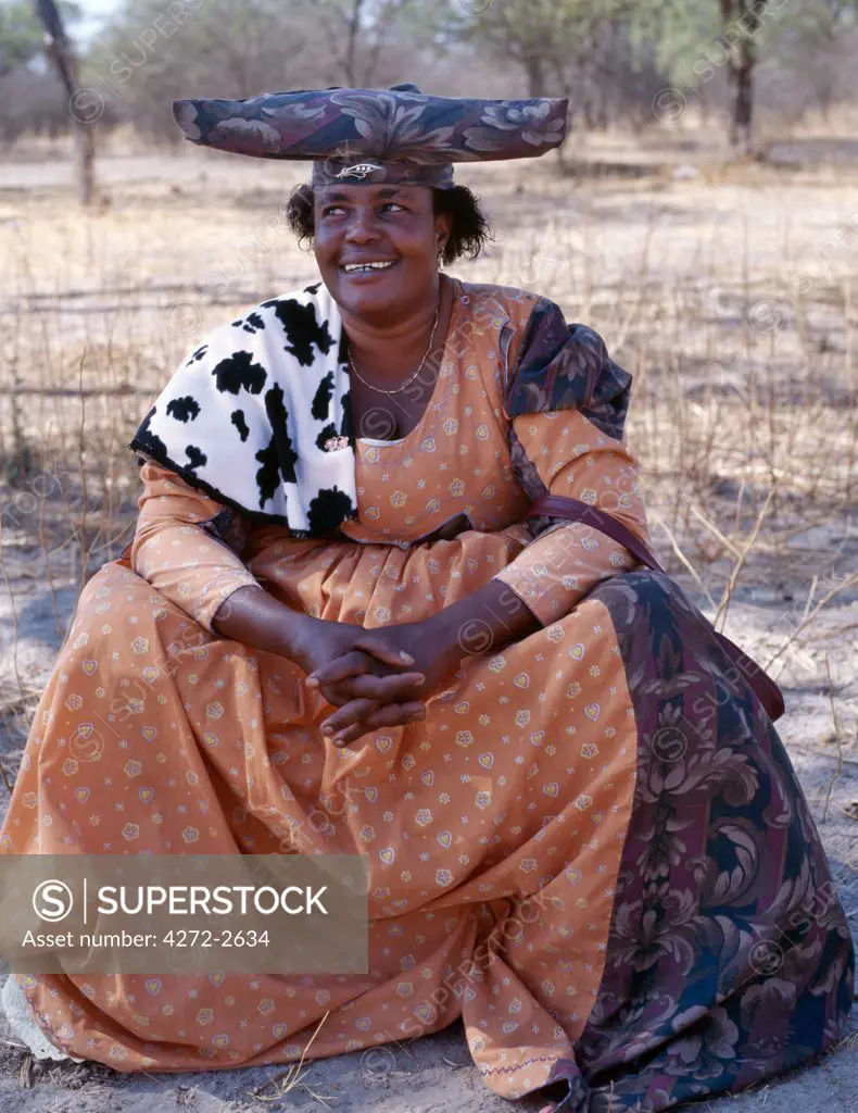 A brightly dressed Herero woman rests in the shade of a tree near her home. The origins of her elaborate dress and unique hat style can be traced back to 19th century German missionaries who took exception to what they considered an immodest form of dress among the tribe. They insisted that Herero women adopt their own impractical style of Victorian era dresses.