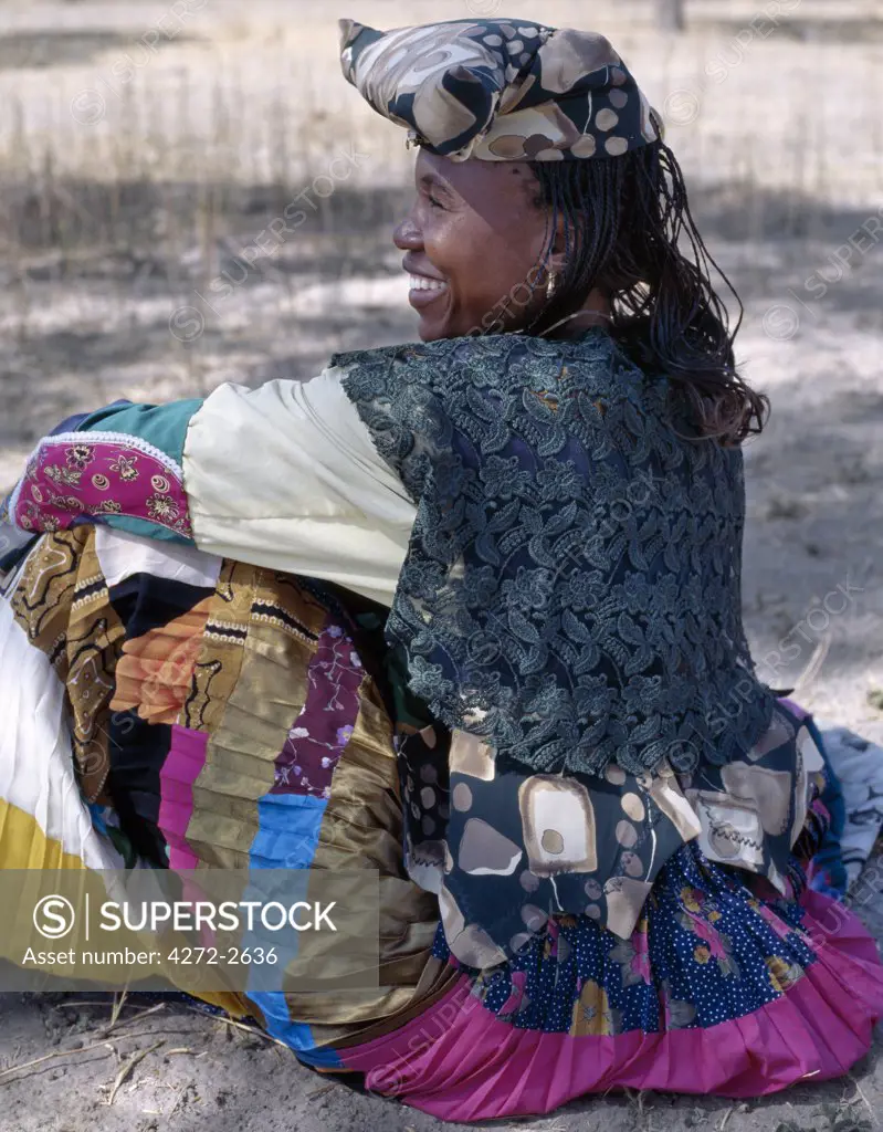A brightly dressed Herero woman rests in the shade of a tree near her home. The origins of her elaborate dress and unique hat style can be traced back to 19th century German missionaries who took exception to what they considered an immodest form of dress among the tribe. They insisted that Herero women adopt their own impractical style of Victorian era dresses.