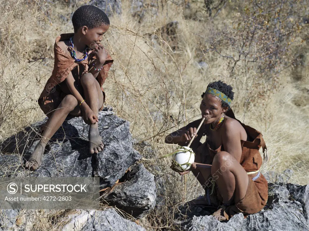 A NS boy watches a woman quench her thirst by sucking water through a hollow reed from a blown ostrich egg. The NS live in the harsh environment of a vast expanse of flat sand and bush scrub country straddling the Namibia Botswana border.