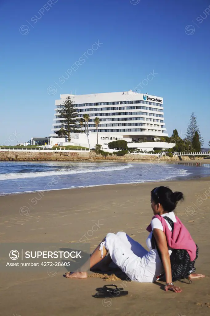Woman relaxing on beach with Beacon Island Hotel in background, Plettenberg Bay, Western Cape, South Africa (MR)