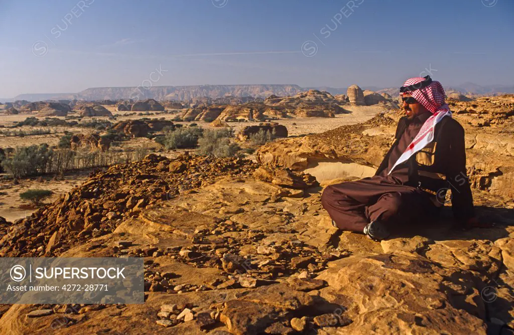 Saudi Arabia, Madinah, nr. Al-Ula, Madain Saleh (aka Hegra). A Saudi man gazes at stark cliffs and rocky outcrops that mark the site of Hegra, once an ancient Nabatean settlement and now a UNESCO World Heritage Site.