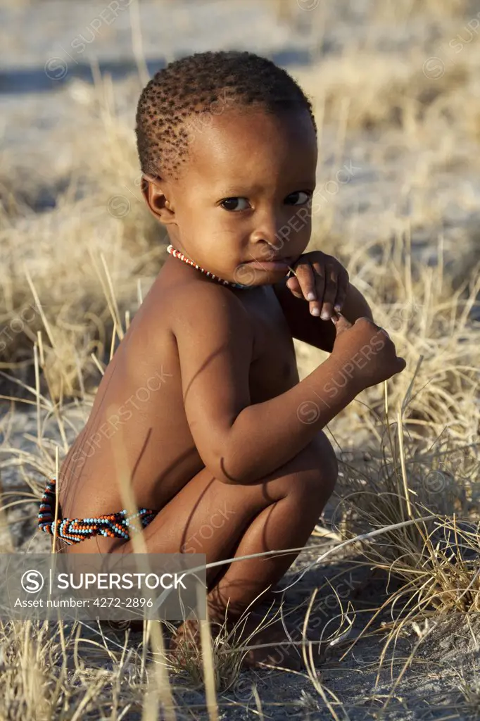 Botswana, Makgadikgadi. A San bushman baby plays in the dry grass, wearing just a traditional belt of beads.
