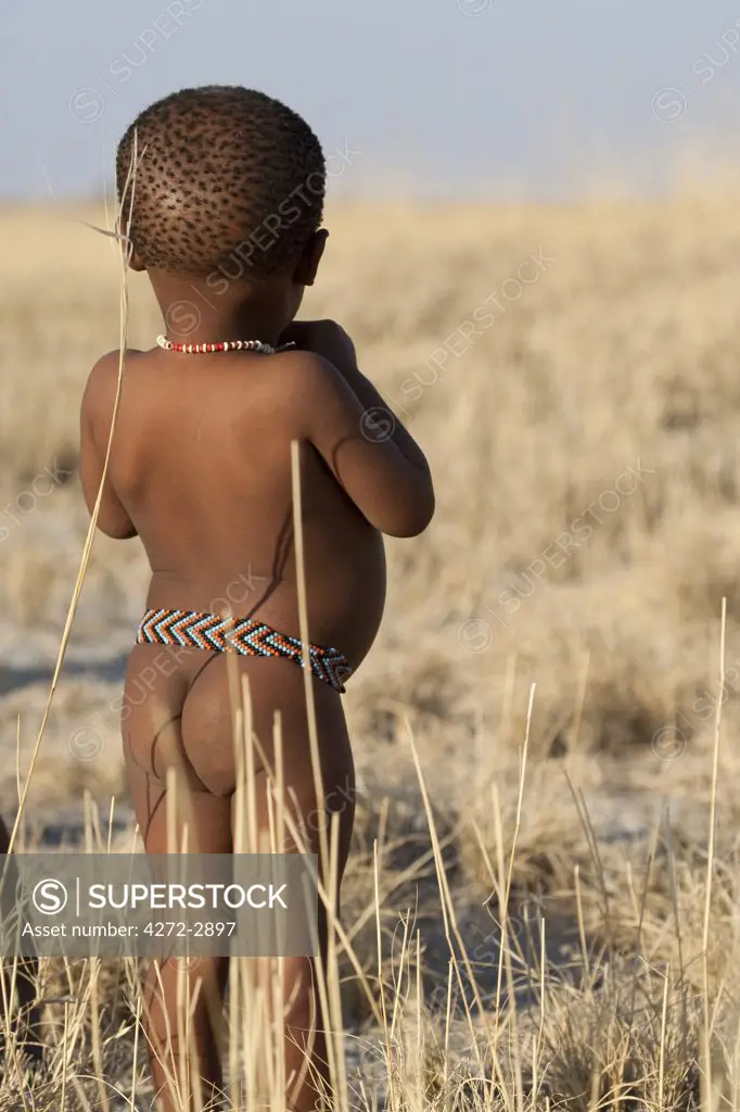 Botswana, Makgadikgadi. A San bushman baby plays in the dry grass, wearing just a traditional belt of beads.