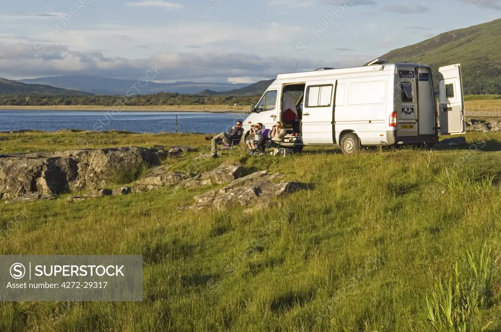 A family on a camping holiday sit outside their campervan on the shore of Loch na Keal on a warm evening enjoying the prolific birdlife