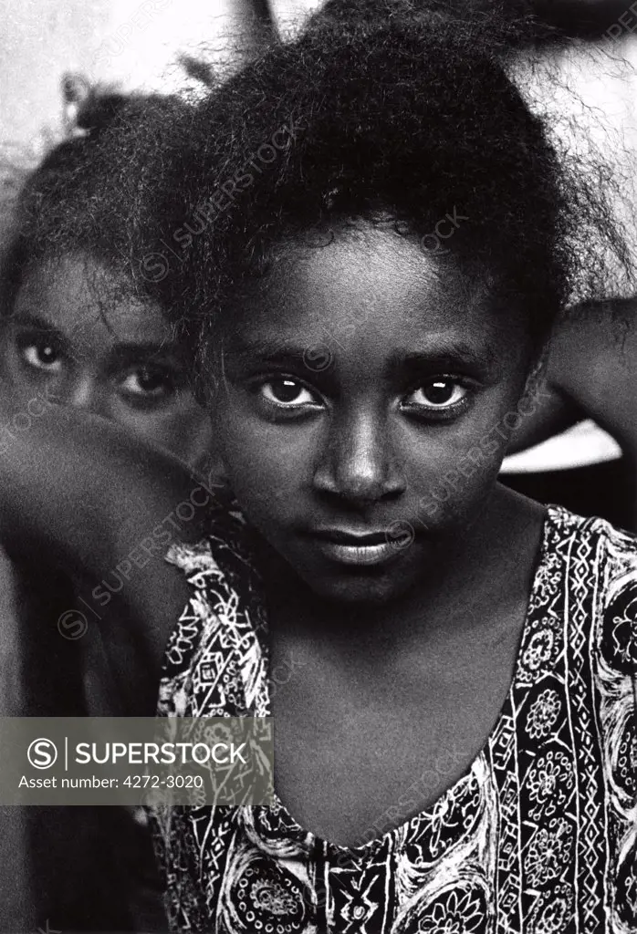 A young Brazilian girl looks directly in the camera, with her sister looking on in the background.