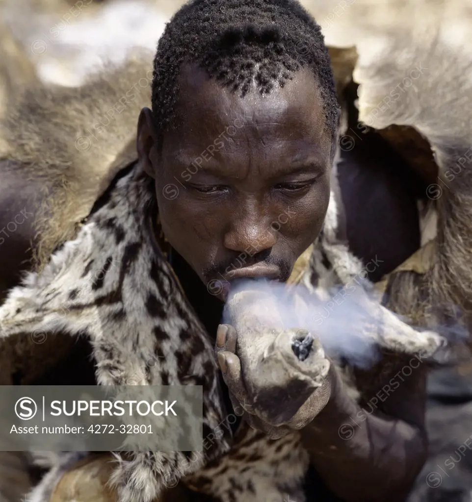 A Hadza hunter wearing a genet cat skin cape smokes cannabis from a crude stone pipe sheathed in leather.The Hadzabe are a thousand-strong community of hunter-gatherers who have lived in the Lake Eyasi basin for centuries.  They are one of only four or five societies in the world that still earn a living primarily from wild resources.