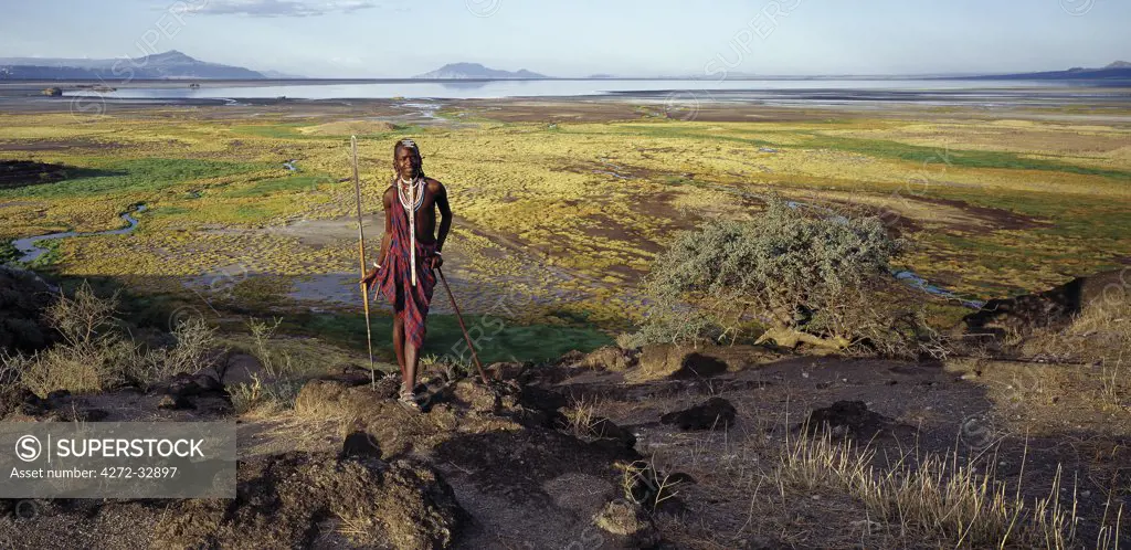 Maasai warrior in front of  Lake Natron in northern Tanzania, one of the most alkaline lakes of the Rift system. As its waters evaporate in the intense heat, sodium sesquicarbonate, known as trona or natron, solidifies to resemble giant coral heads in brightly coloured water. The pastoral Maasai graze their cattle along its southern shores where the Enkare Sero River provides welcome pasture.