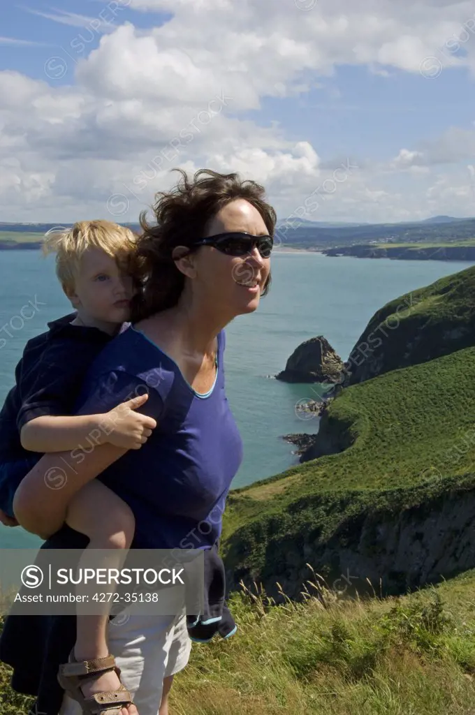 UK, Wales, Pembrokeshire. A mother carries her young son along the Dinas Head section of the Pembrokeshire Coastal Path with views towards Needle Rock and Newport Bay beyond. (MR).