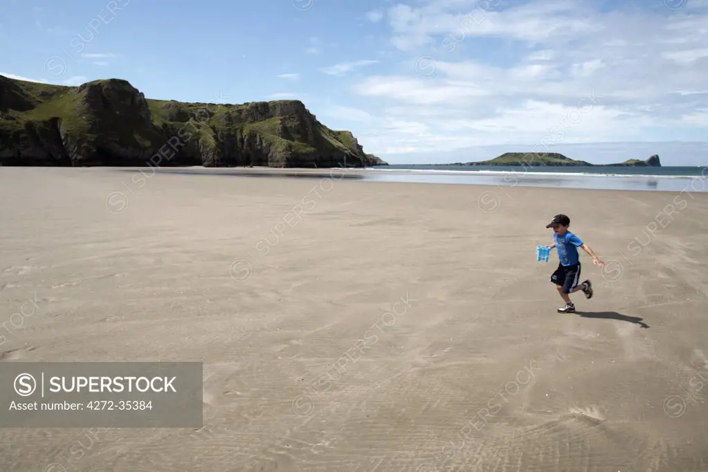 Wales, Gower. Young boy runs across the empty sands of Rhossili Bay at low tide, carrying a bucket of water. (MR)
