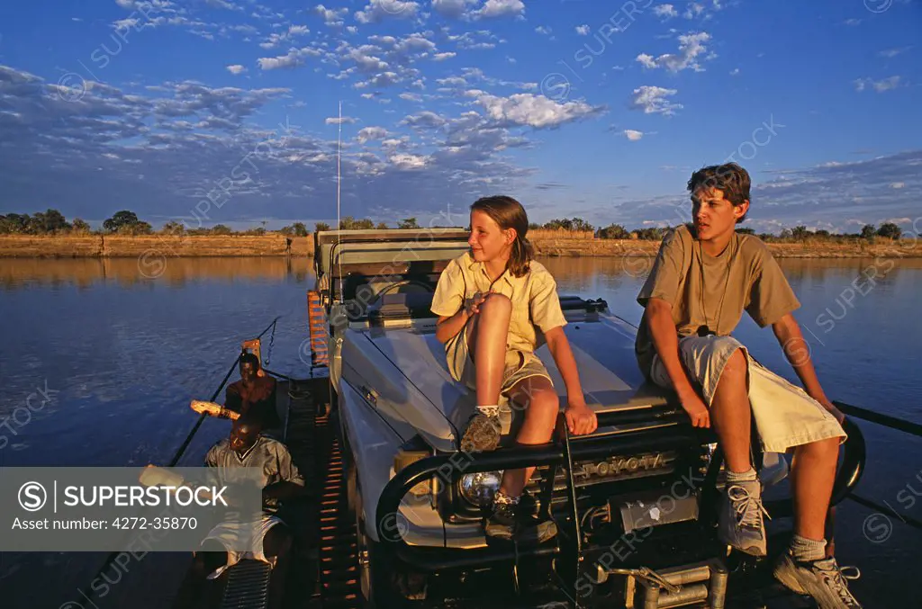 Zambia, South Luangwa National Park. Children sit on the bonnet of the landcruiser during a pontoon crossing of the Luangwa River on a family safari.