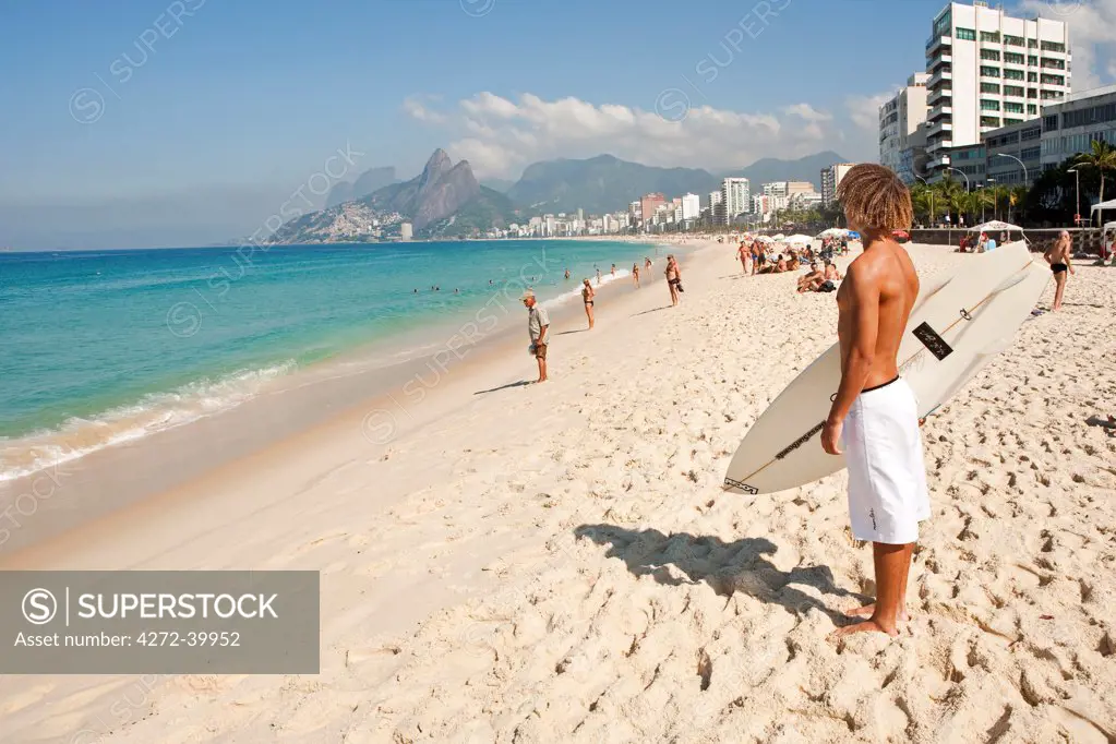 South America, Rio de Janeiro, Rio de Janeiro city, Ipanema, surfer with a surf board looking out over the Atlantic from the Arpoador on Ipanema beach with the Dois Irmaos mountains in the distance