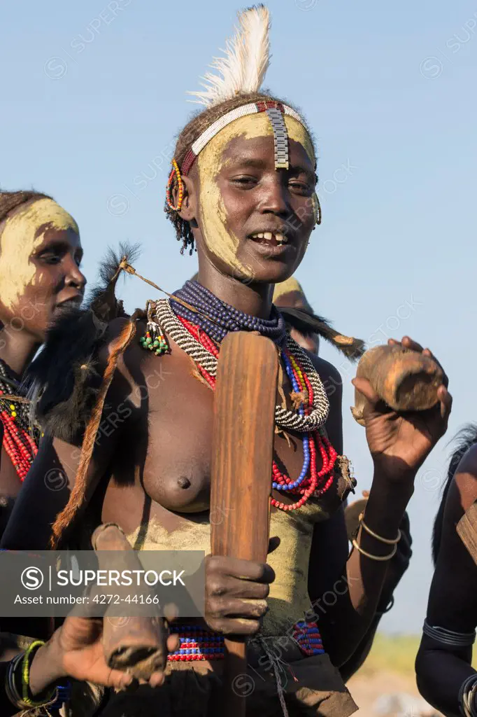 A Dassanech woman dressed in ceremonial regalia, holding a wooden baton and a cowhorn container, participates in a Dimi dance, Ethiopia