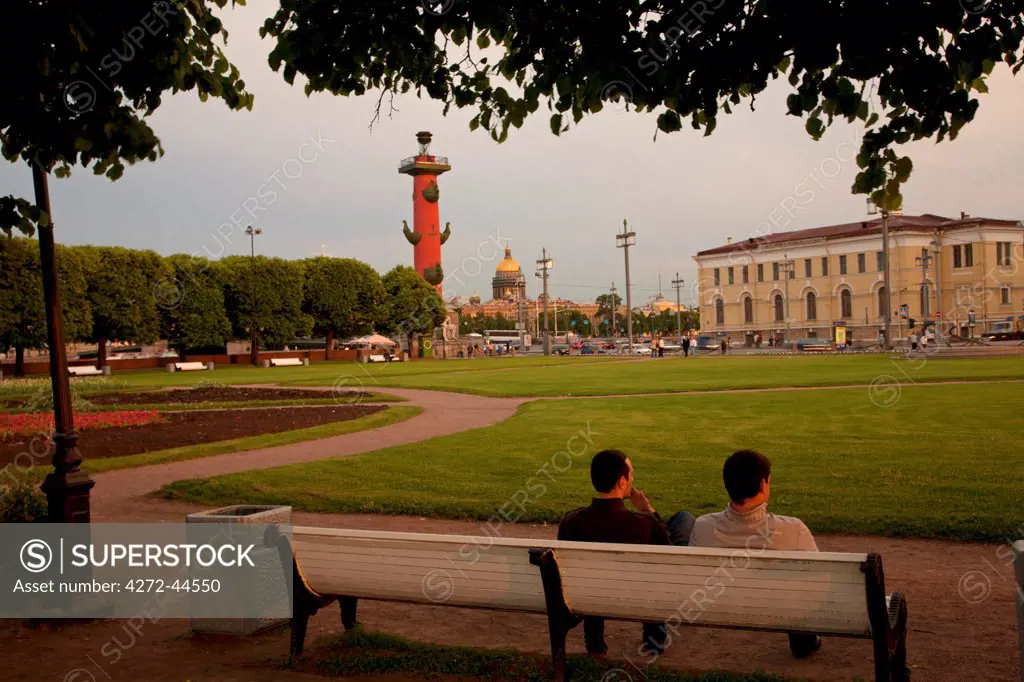 Russia, St.Petersburg. Two men enjoying the atmosphere in a garden during the White Nights on Vassilevski island facing one of the Rostral Columns with St.Isaac Cathedral in the background