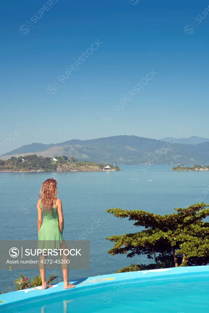 Brazil, Rio de Janeiro, Parati, a woman in a green dress looks out over the Bay of Parati from the edge of a swimming pool MR