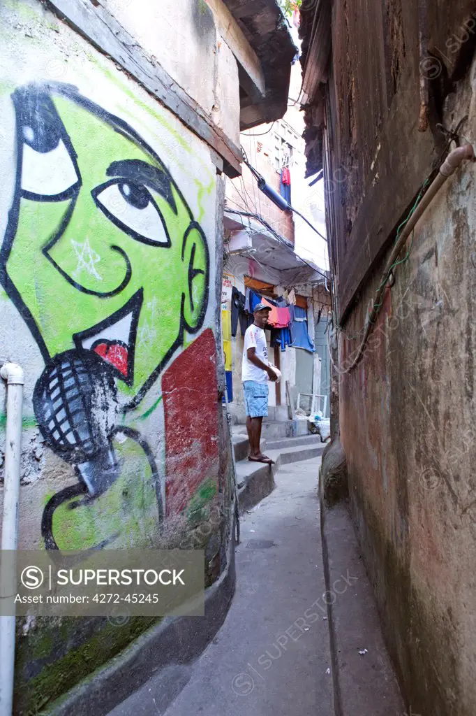 South America, Rio de Janeiro, Rio de Janeiro city, a local smiling at the camera in a narrow alley with colourful graffiti running between breeze block houses in the Dona Marta favela, Santa Marta community