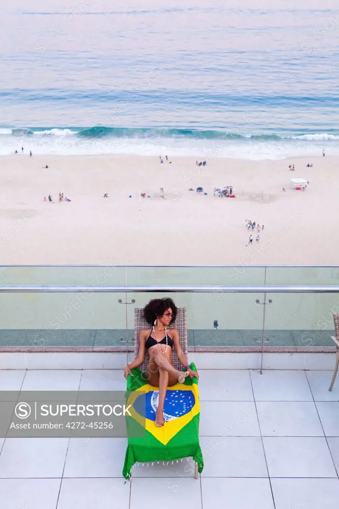 South America, Brazil, Rio de Janeiro, a model in a black bikini sunbathes on a shawl based on the Brazilian flag with Copacabana Beach behind her MR