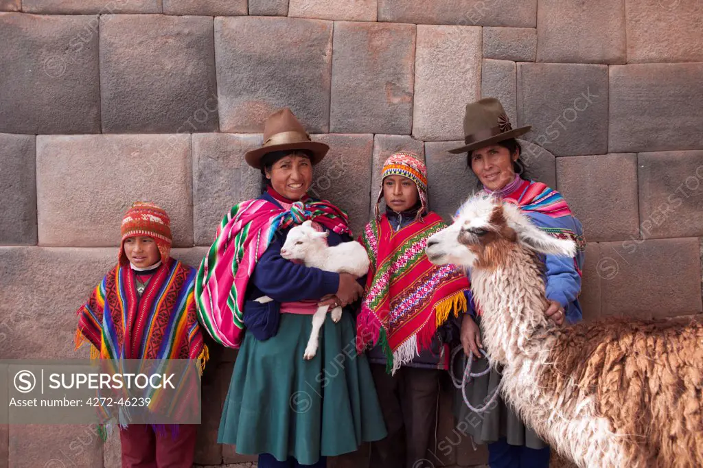 South America, Peru, Cusco. Quechua people standing in front of an Inca wall, holding a lamb and a llama and wearing traditional clothing including a bowler hat, liclla, chullo and poncho    while talking on a cell phone in the UNESCO World Heritage listed former Inca capital of Cusco, MR,