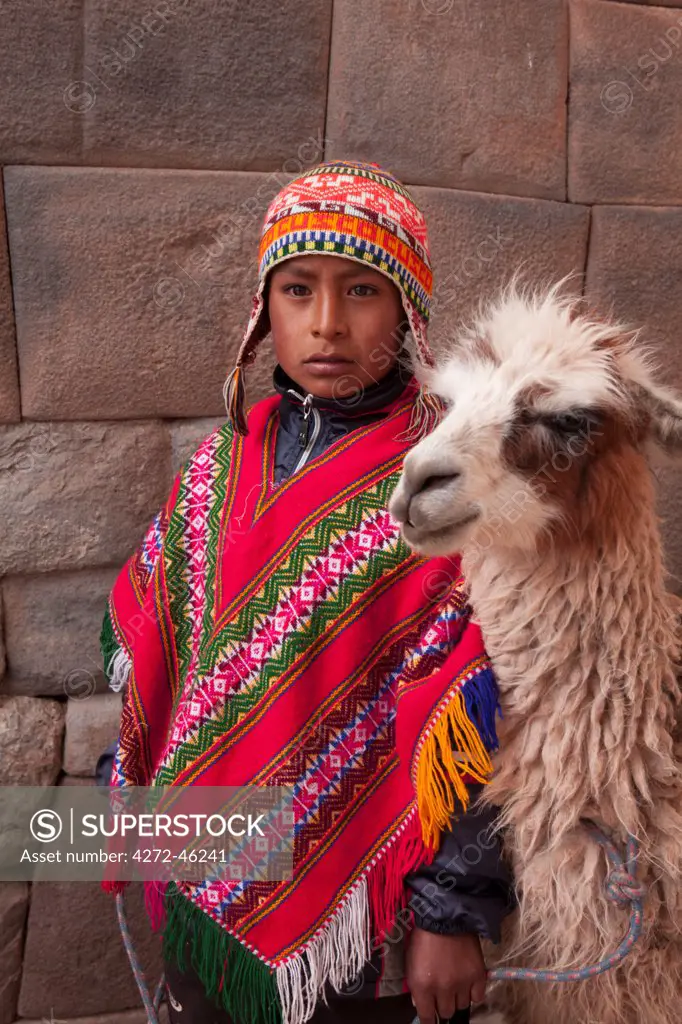 South America, Peru, Cusco. A Quechua boy in a poncho and a chullo woollen cap with a Llama standing in front of an Inca wall in the UNESCO World Heritage listed former Inca capital of Cusco