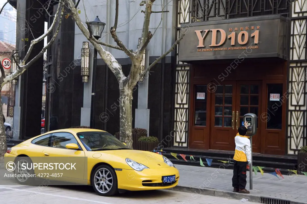 China, Shanghai. French Concession area - a young boy putting coins in the parking meter next to a yellow Porshe.