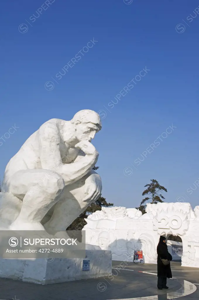 China, Northeast China, Heilongjiang Province, Harbin City. Snow and Ice Sculpture Festival at Sun Island Park. Visitors standing beneath a thinking man sculpture.
