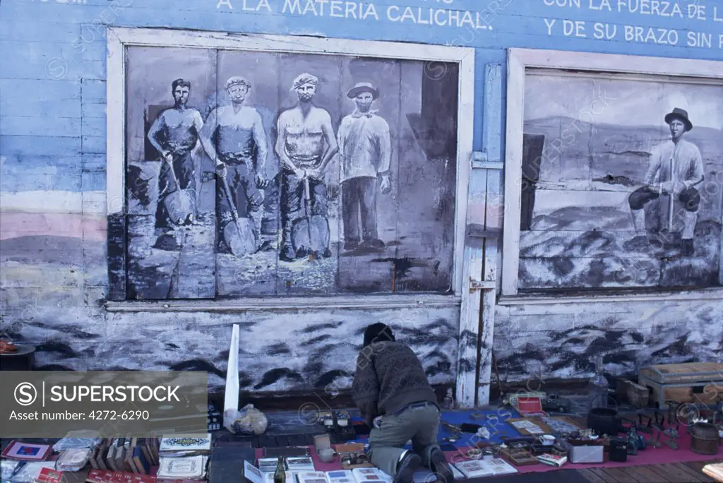 A street trader lays out his wares, artefacts from the nineteenth century nitrate boom, beneath street art depicting the nitrate miners. Situated on the coast at the foot of the 800 metre coastal cordillera, Iquique is the capital of Region 1.  During the nitrate boom in the 19th century Iquique became Chiles richest city.