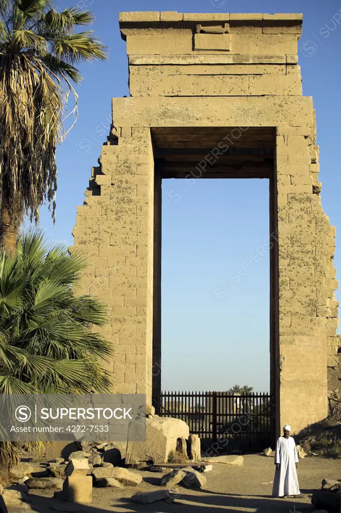 A egyptian man in traditional dress stands in front of a colossal gateway in the Temple of Karnak, Luxor.