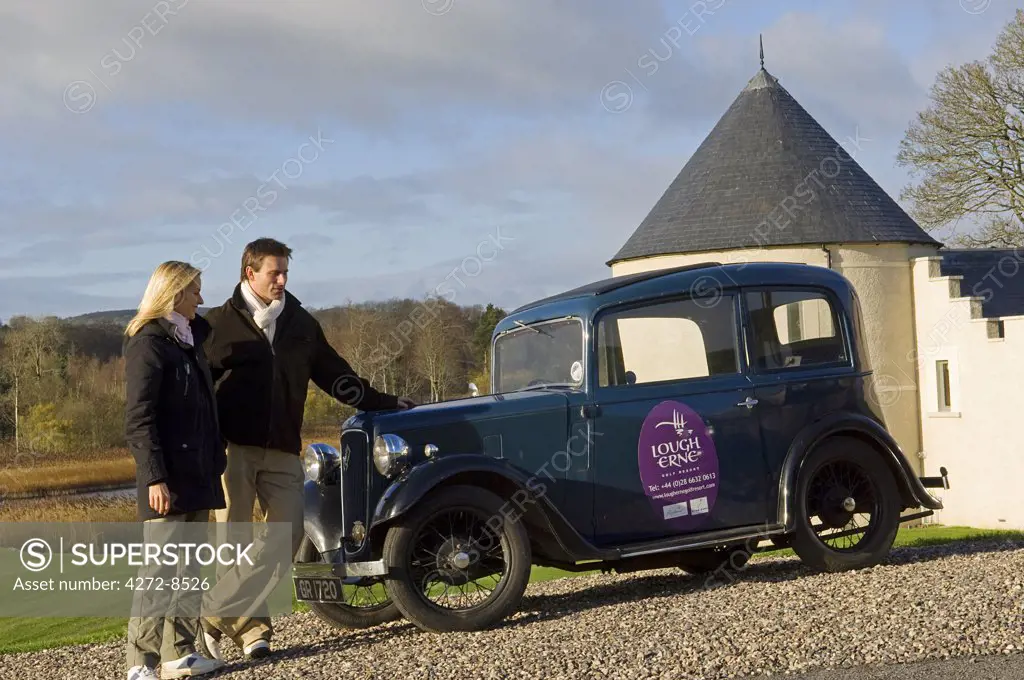 UK, Northern Ireland, Fermanagh, Enniskillen. A couple of guests stand beside a vintage car at the entrance to Lough Erne Golf Resort (MR).