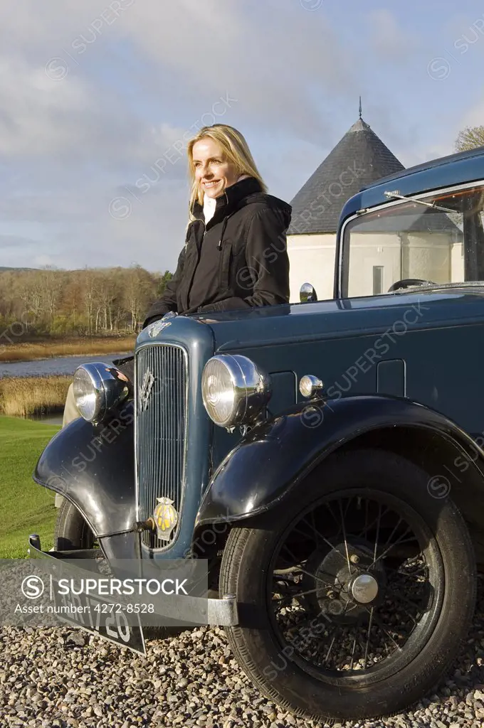 UK, Northern Ireland, Fermanagh, Enniskillen. A female guest stands beside a vintage car at the entrance to Lough Erne Golf Resort (MR).