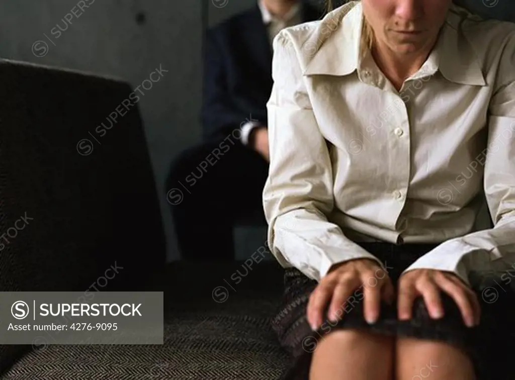 Woman sitting with hands on knees, therapist behind her, cropped