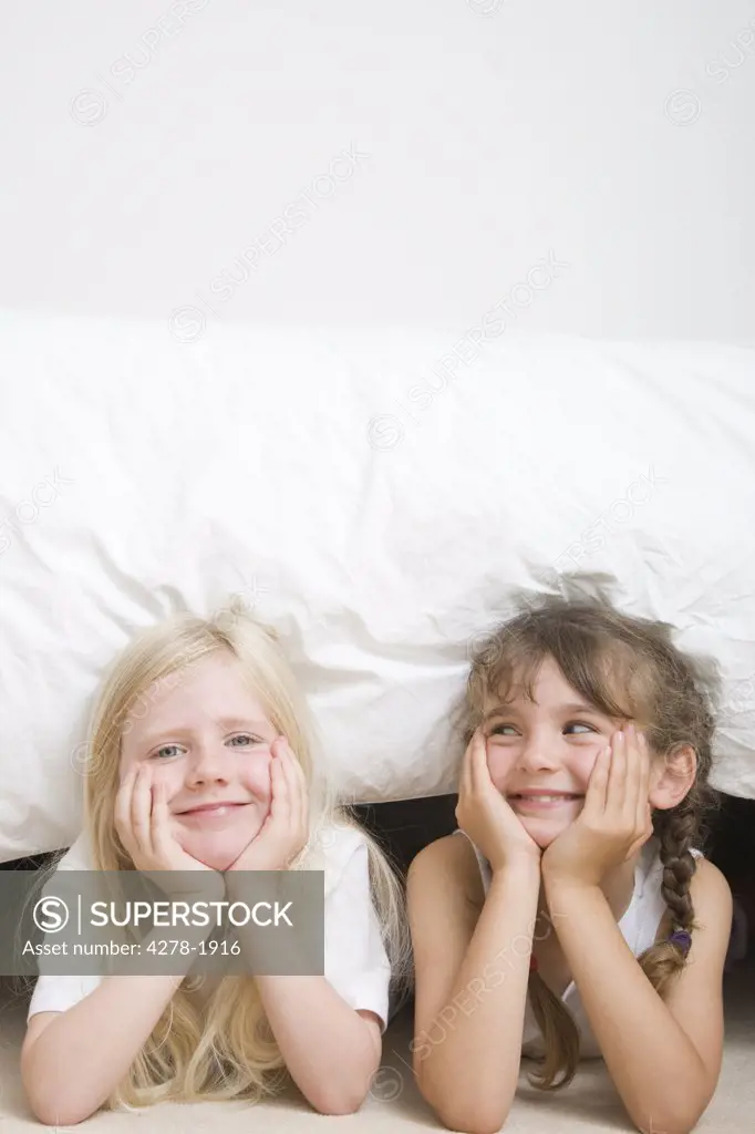 Two young girls with chin resting on hands under bed smiling