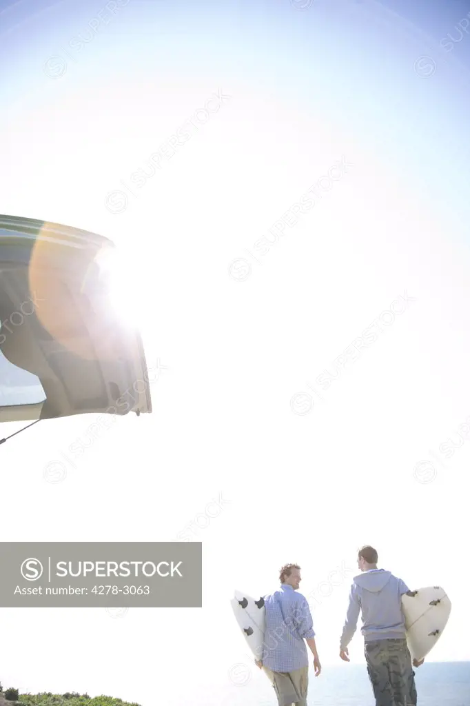 Two young men walking away from car holding surfboards