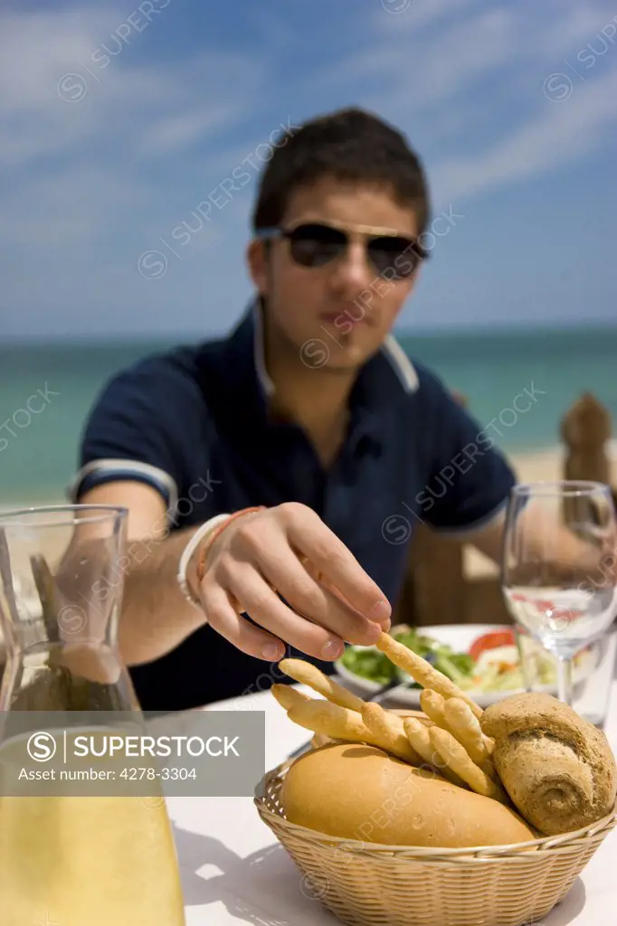 Young man eating al fresco by the sea picking up a breadstick from bread basket