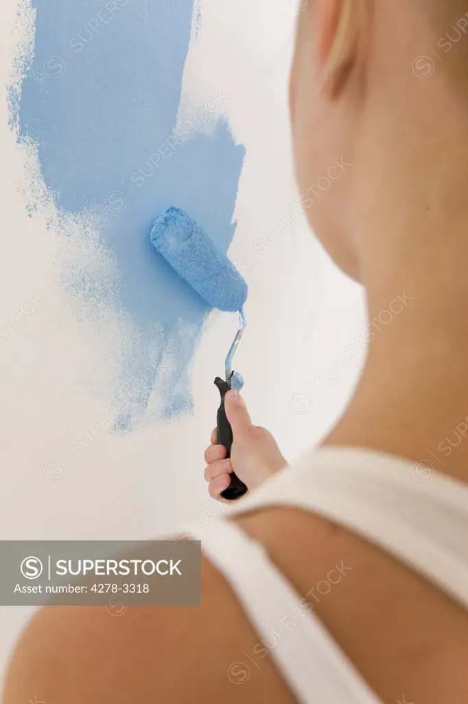 Back view of woman holding paint brush and painting a white wall with blue paint