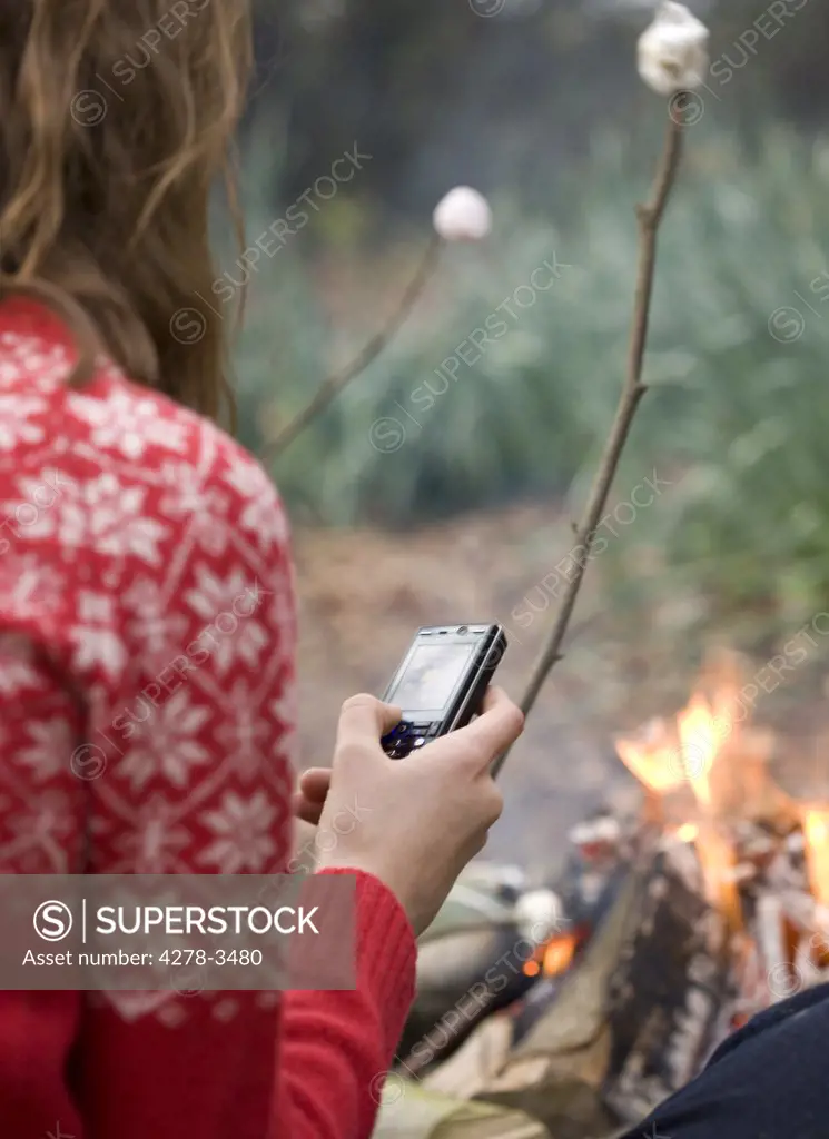 Back view of teenaged girl roasting marshmallow over campfire using cell phone