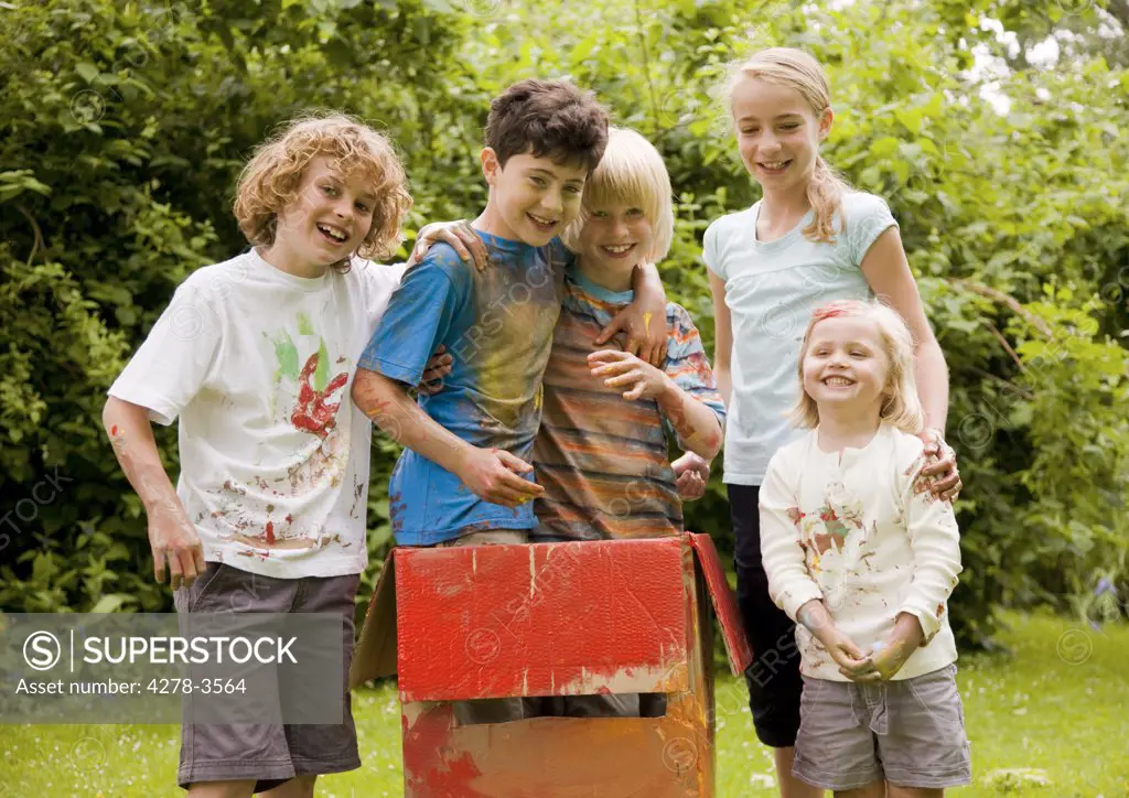 Children covered in watercolor paint in a garden, two of them standing in a cardboard box