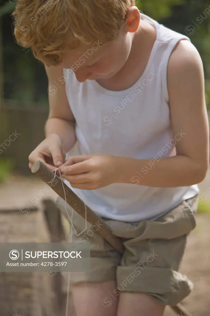 Young boy holding wooden stick between legs tying a knot with a string