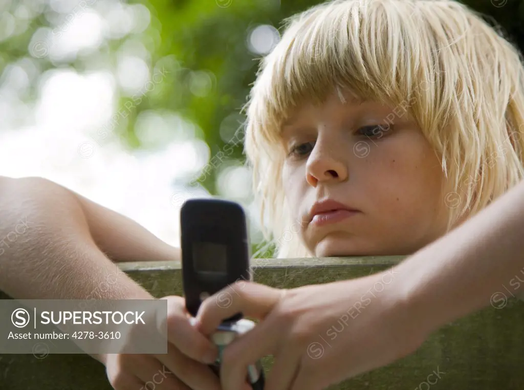 Young boy with arms over wooden fence holding and looking at cell phone