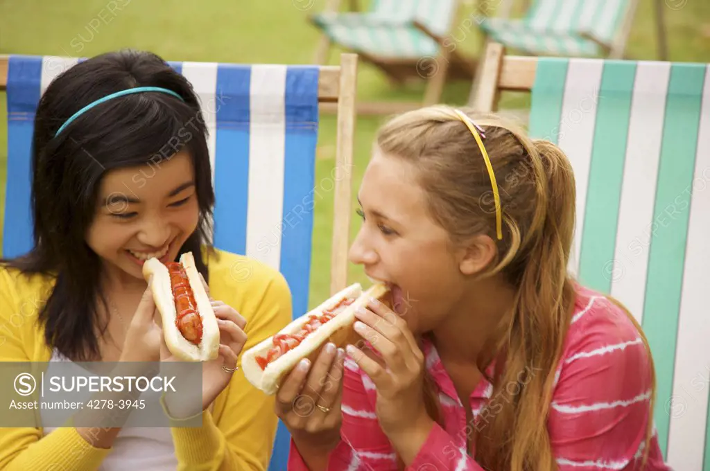 Two teenaged girls sitting on deck chairs in a London park eating hot dogs and laughing
