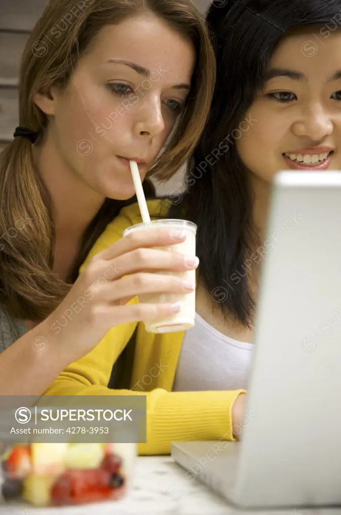 Two teenaged girls sitting in front of laptop computer typing and drinking milk-shake