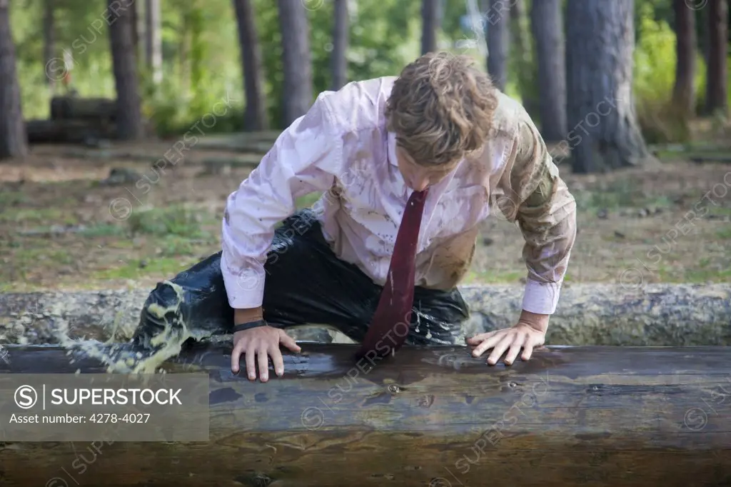 Businessman at obstacle course clambering over a wooden beam soaked in muddy water