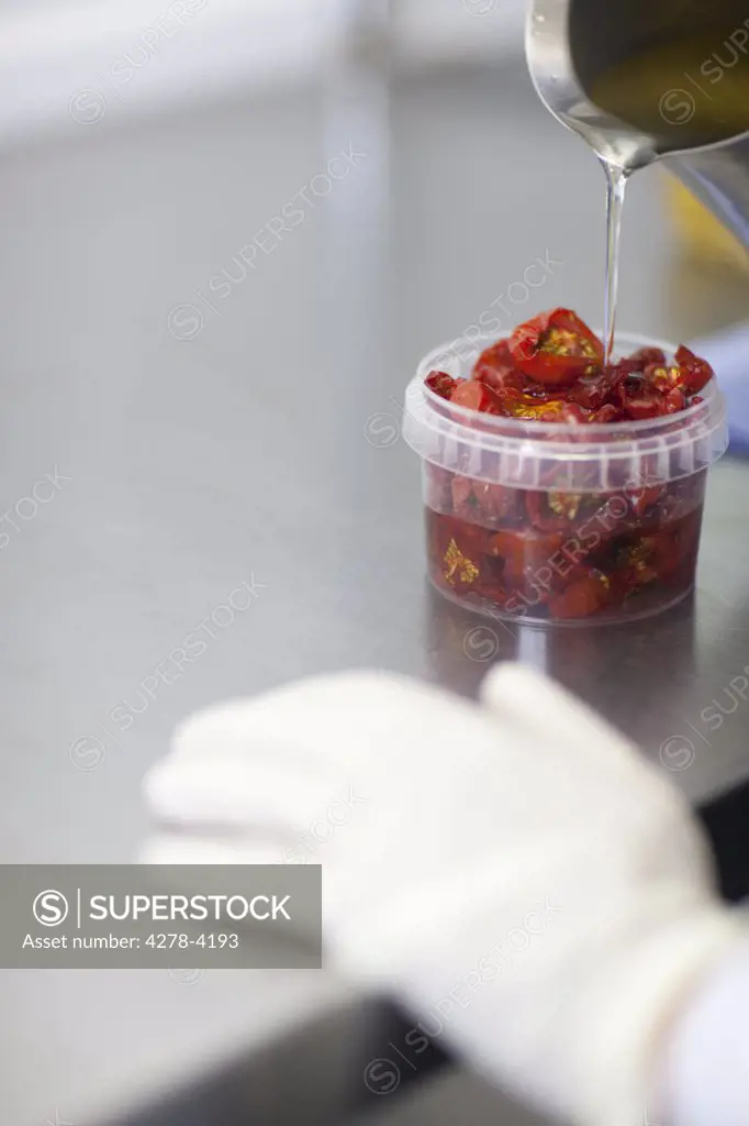 Close up of a worker hand pouring oil in a plastic container filled with sun dried tomatoes