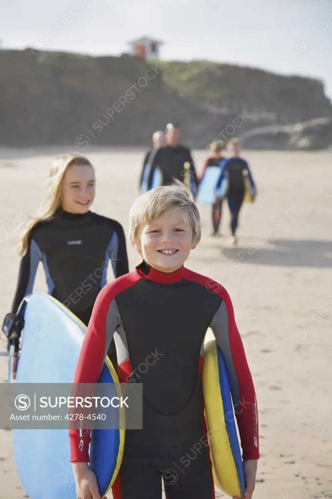 Boy and girl carrying surfboards on a beach followed by people