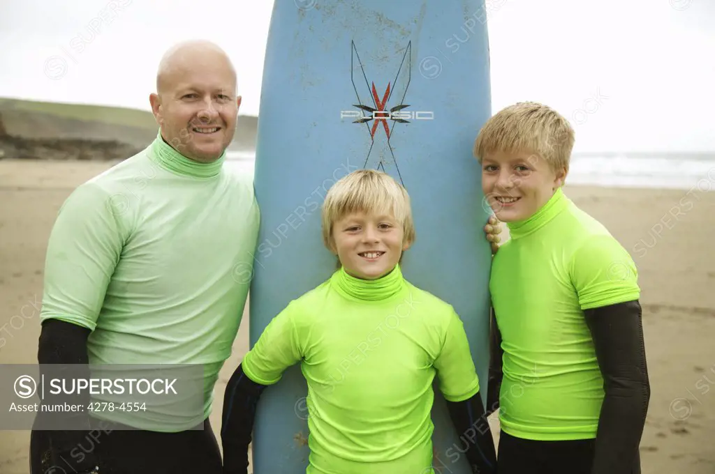 Man and two boys wearing surfing wetsuits standing around an upright surfboard on a beach smiling