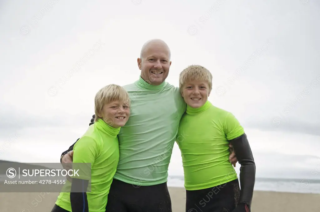 Man and two boys wearing surfing wetsuits standing on a beach smiling