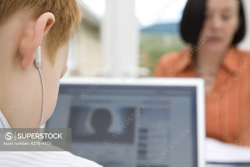 Back view of a boy using a laptop and wearing earphones sitting opposite woman