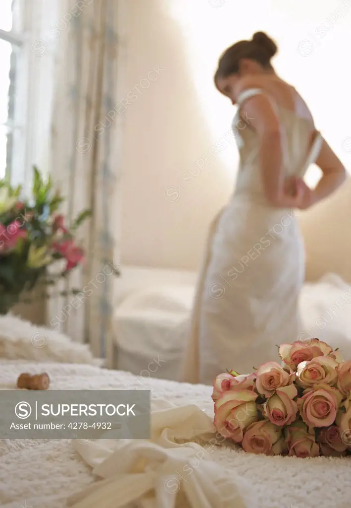 Close up of a bouquet of roses and bridal gloves lying on a bed with bride undressing