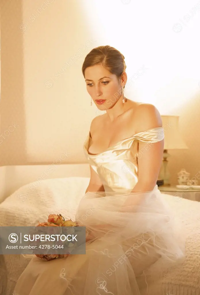 Bride in a white wedding gown sitting on a bed holding a bouquet of roses