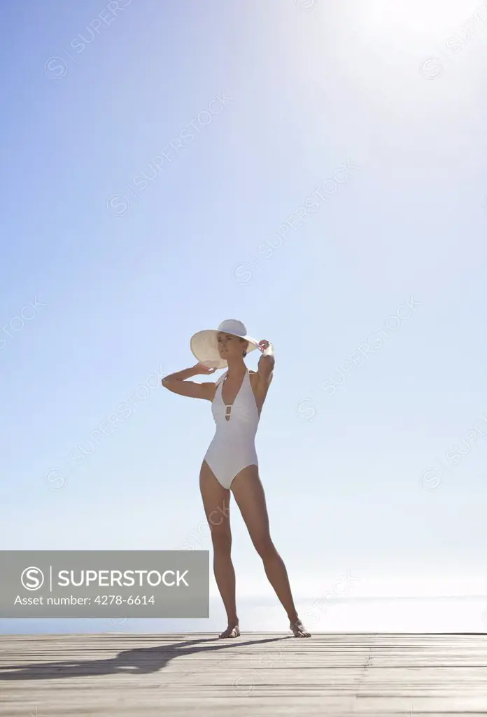 Woman in white hat and swimsuit standing on a sun deck holding the brim of her hat