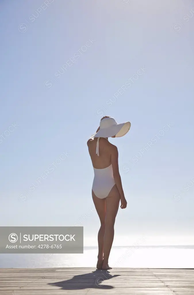 Woman in white swimsuit standing on a sun deck holding her hat